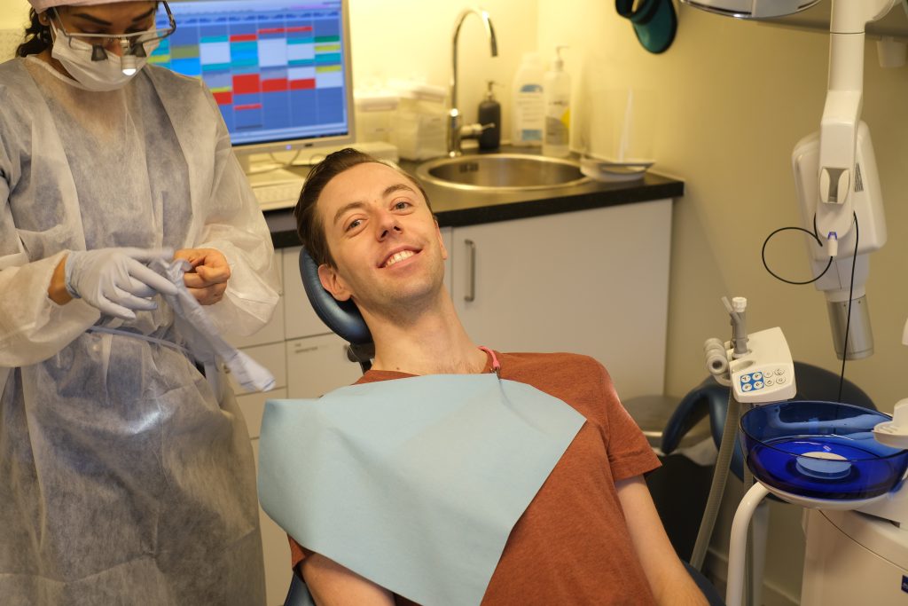 Mann sitter i tannlegestol smilende mot kamera. En tannlege står bak og forbereder seg på å sjekke tennene på mannen ved å ta på seg hansker