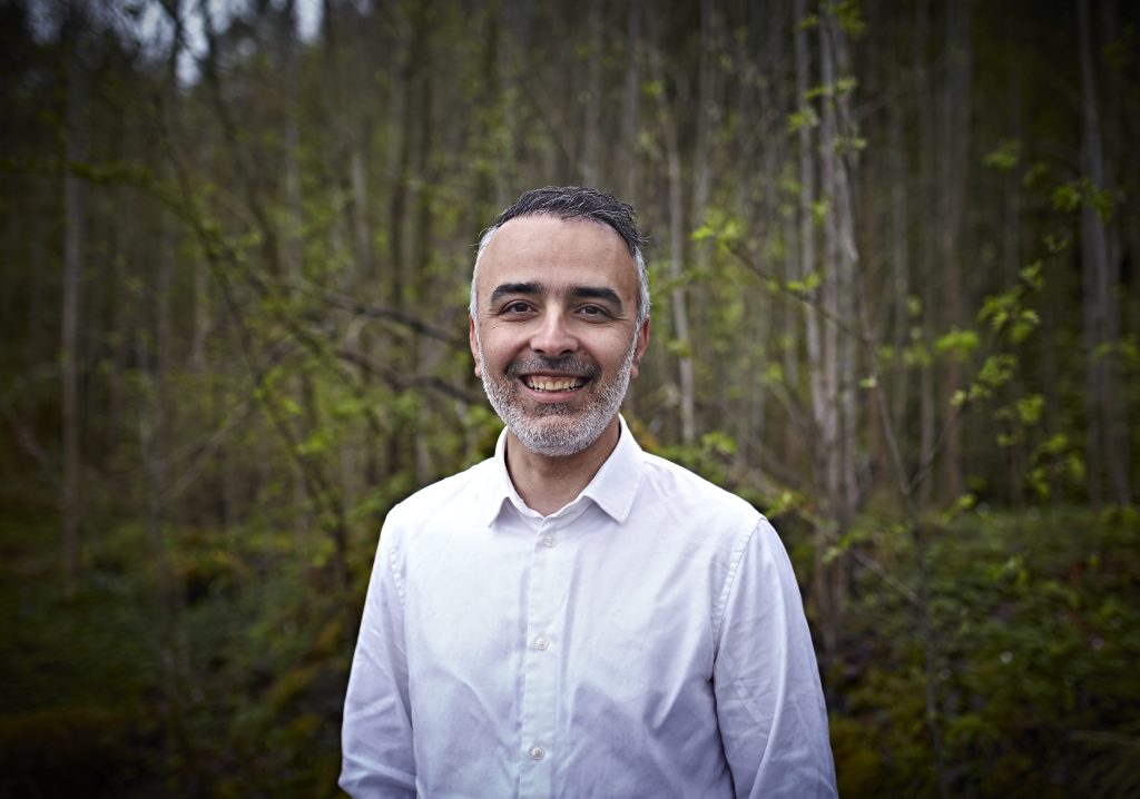 Bilde i profil av Barham Azadi i Hvit skjorte. Barham smiler mot kamera, i med en mosegrodd skog i bakgrunnen.