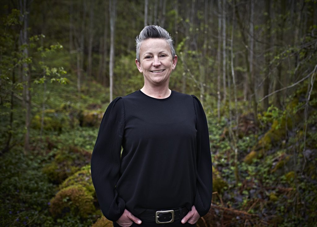Bilde i profil av Åse Heien i en sort topp med kort hår. Åse smiler mot kamera, i med en mosegrodd skog i bakgrunnen.